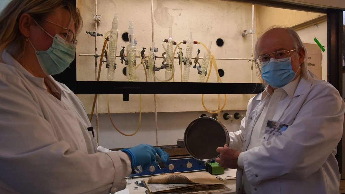 Zwei Personen in Laborkitteln, mit Handschuhen und Masken zeigen eine Staubprobe in einer Messkammer. Im hintergrund sind Laborgerätschaften zu erkennen